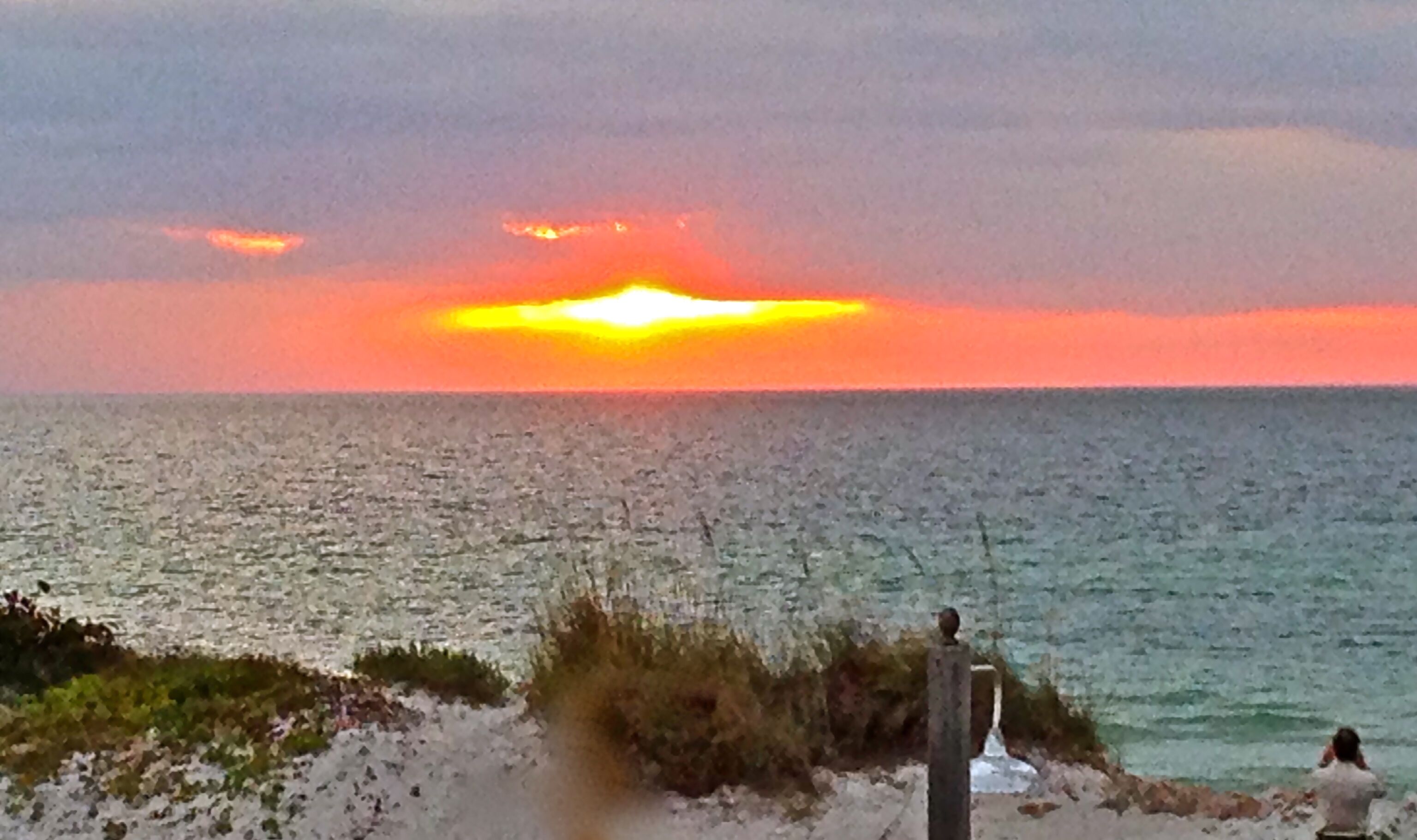 A sunset over a calm sea with a rocky shoreline and a statue.