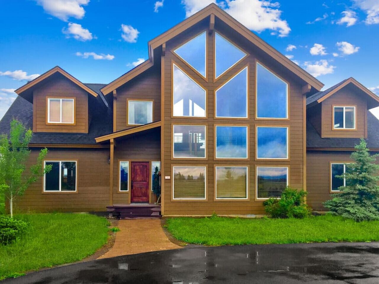 Une maison moderne à deux étages, dotée d’une grande baie vitrée centrale, d’une porte rouge et d’une pelouse bien entretenue.
