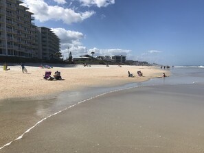 Une plage avec un rivage sablonneux, quelques personnes et des bâtiments en arrière-plan.
