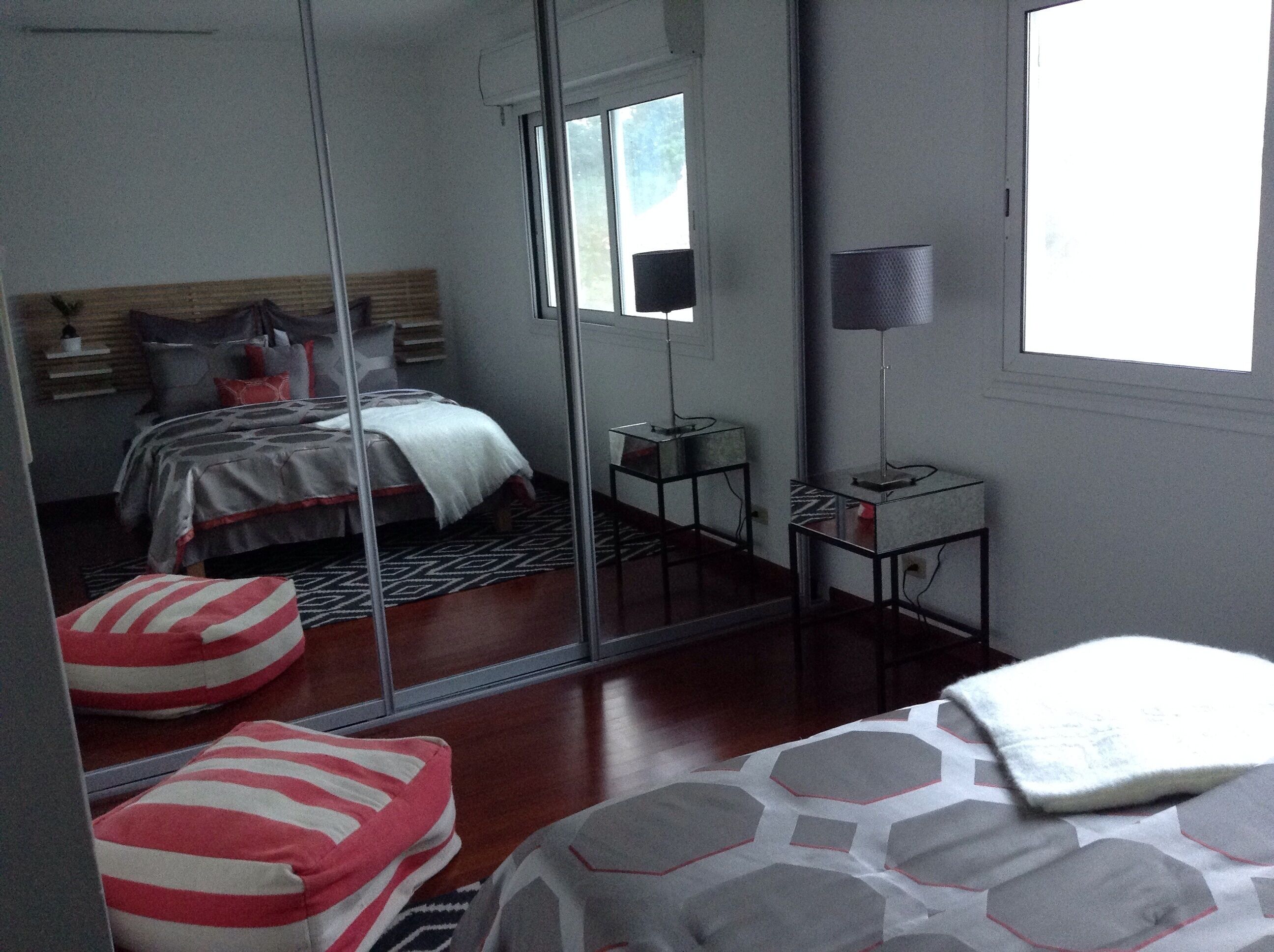 Een slaapkamer met een bed, nachtkastje, lamp, spiegel en raam.