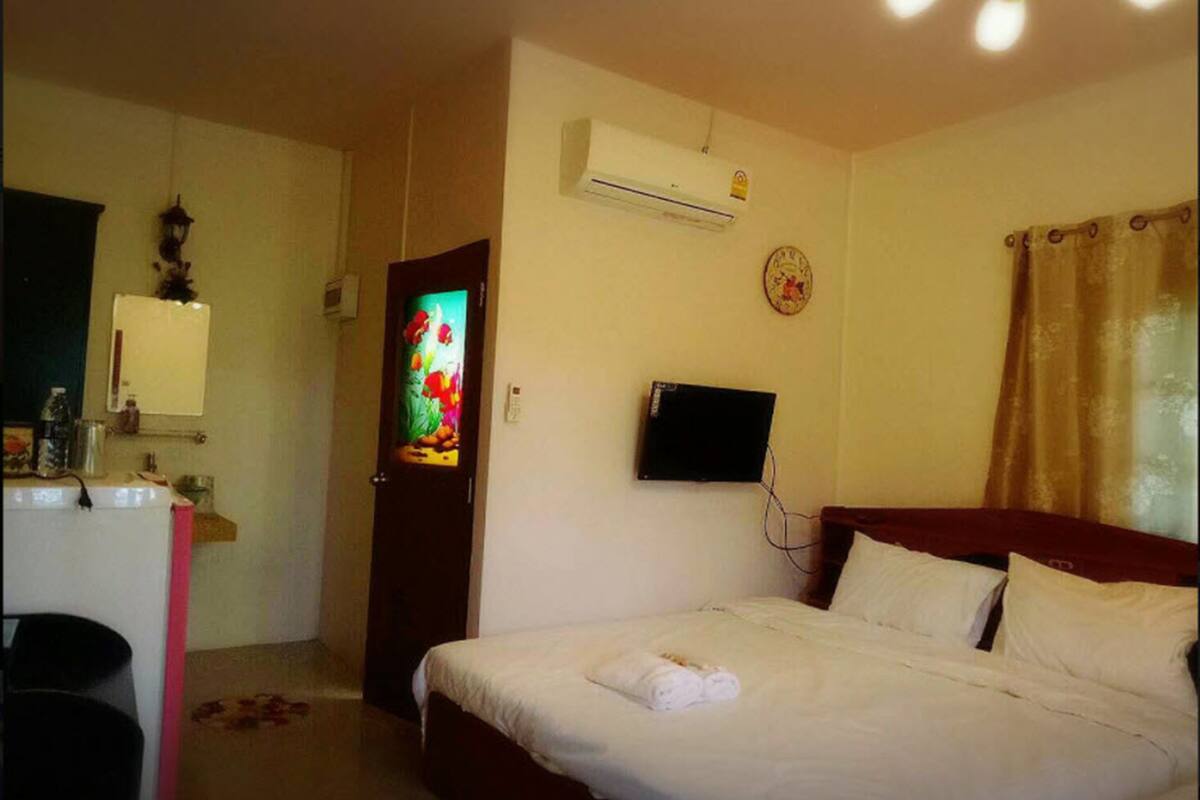 Una habitación de hotel con cama, televisor montado en la pared, aire acondicionado, un pequeño escritorio con espejo y una puerta decorada con una obra de arte colorida.