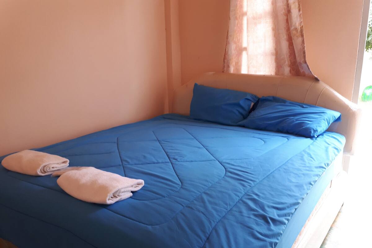Una cama con ropa de cama y almohadas azules, una toalla doblada y una ventana con cortinas.