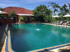 A swimming pool with lounge chairs and umbrellas, surrounded by palm trees and a building with a red roof. - Heaven Hill Pool Villa Pattaya (Pattaya)