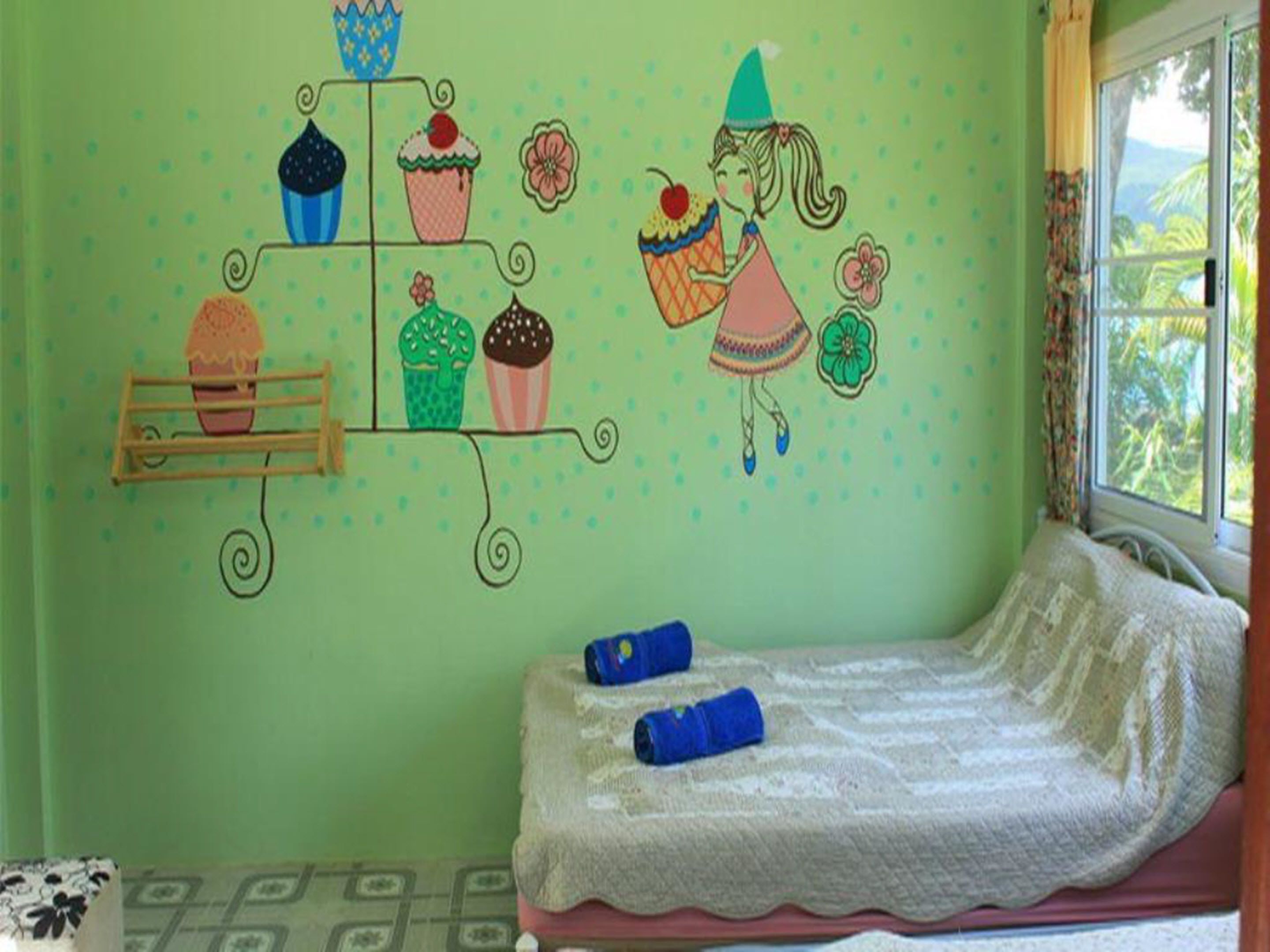 A room with a bed, a window with a view of greenery, and a wall decorated with cupcake illustrations.