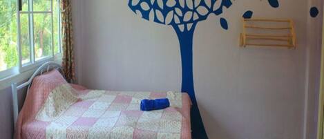 A room with two beds, a window with curtains, a wall mural of a tree with leaves and a bird, and a wooden rack.