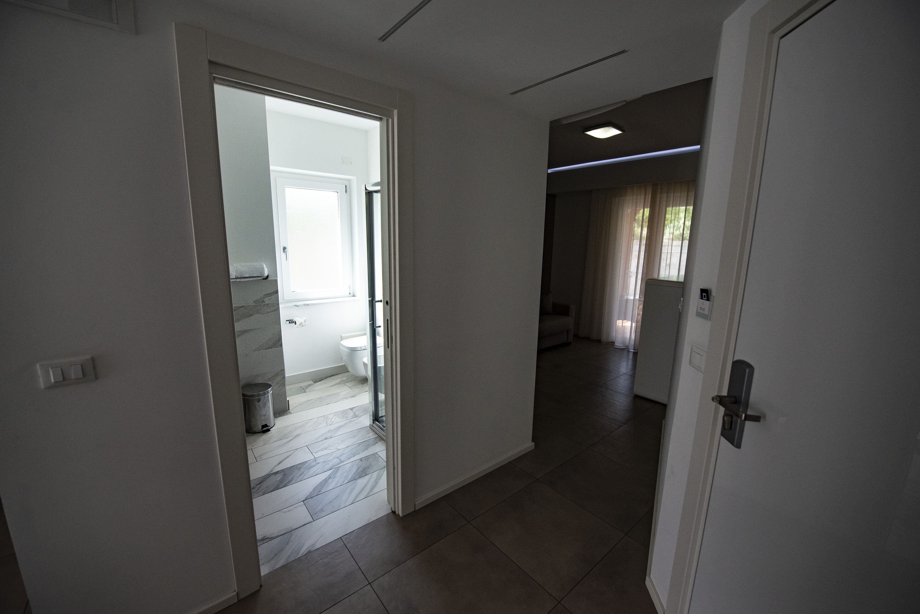 A hallway with a bathroom visible through an open door, featuring a toilet and a trash can.