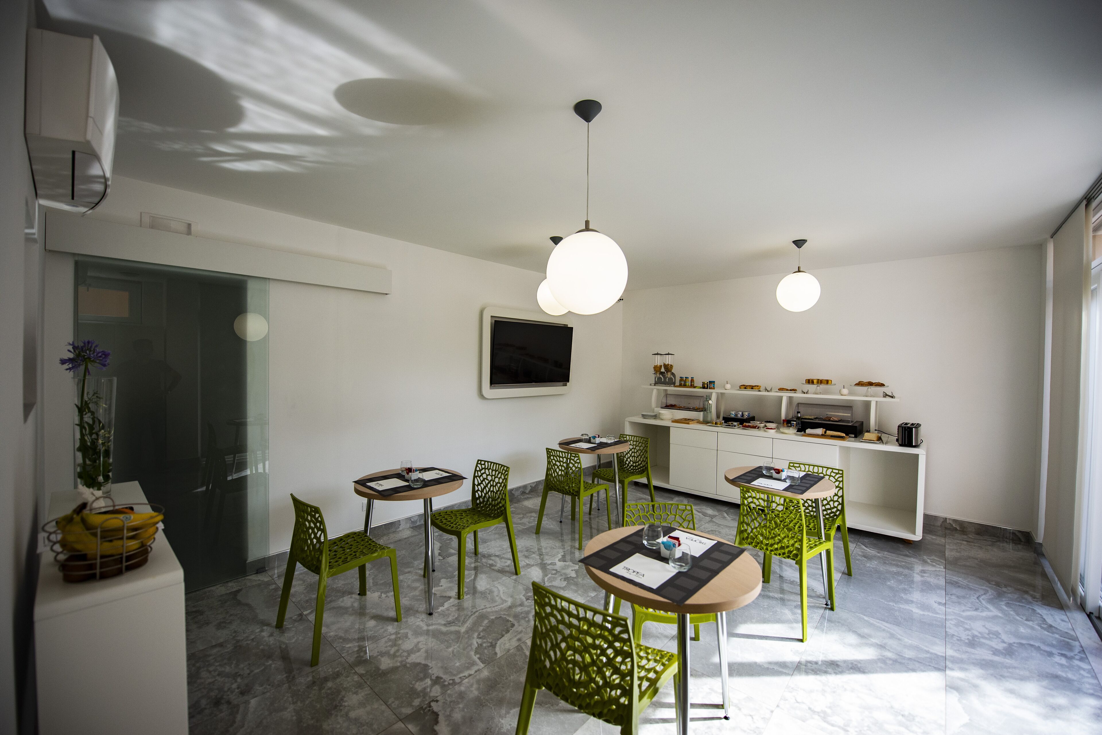 A modern dining area with green chairs and round tables, a kitchen area with white cabinets, and a flat-screen TV mounted on the wall.