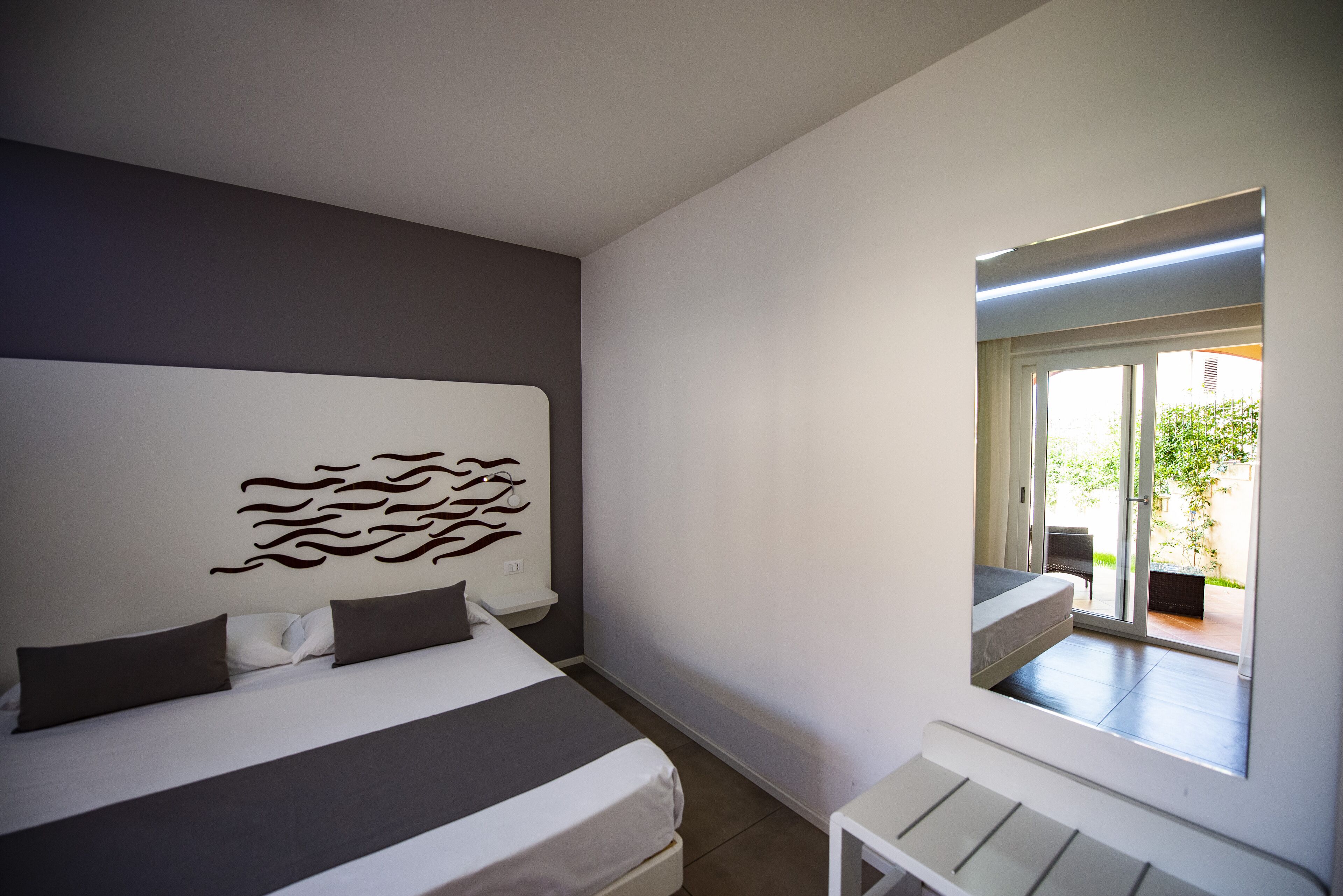 A modern bedroom with a large bed, a headboard with a decorative panel, a small bedside table, and a window with a view of the outdoors.
