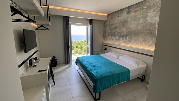 A modern hotel room with a bed, a desk, a chair, a flat-screen TV, and a view of the sea. - Aether Suites Tropea (Tropea)