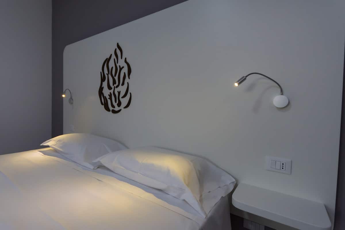 Un lit bien fait, avec du linge de lit blanc, un luminaire fixé au mur et une prise de courant.