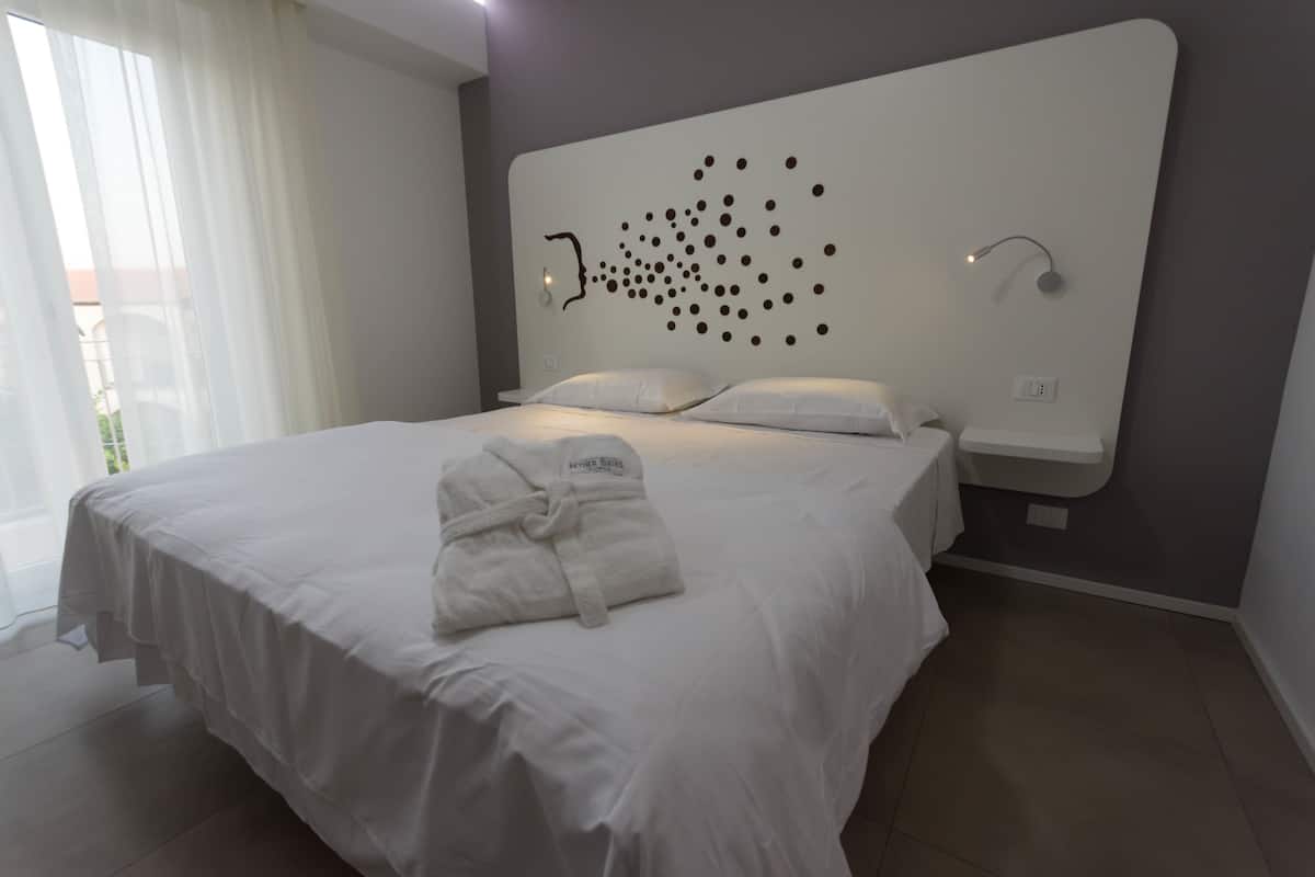 Une chambre d’hôtel moderne avec un grand lit, du linge de lit blanc, une tête de lit ornée d’un motif à pois original, un peignoir posé sur le lit et une petite table de chevet.