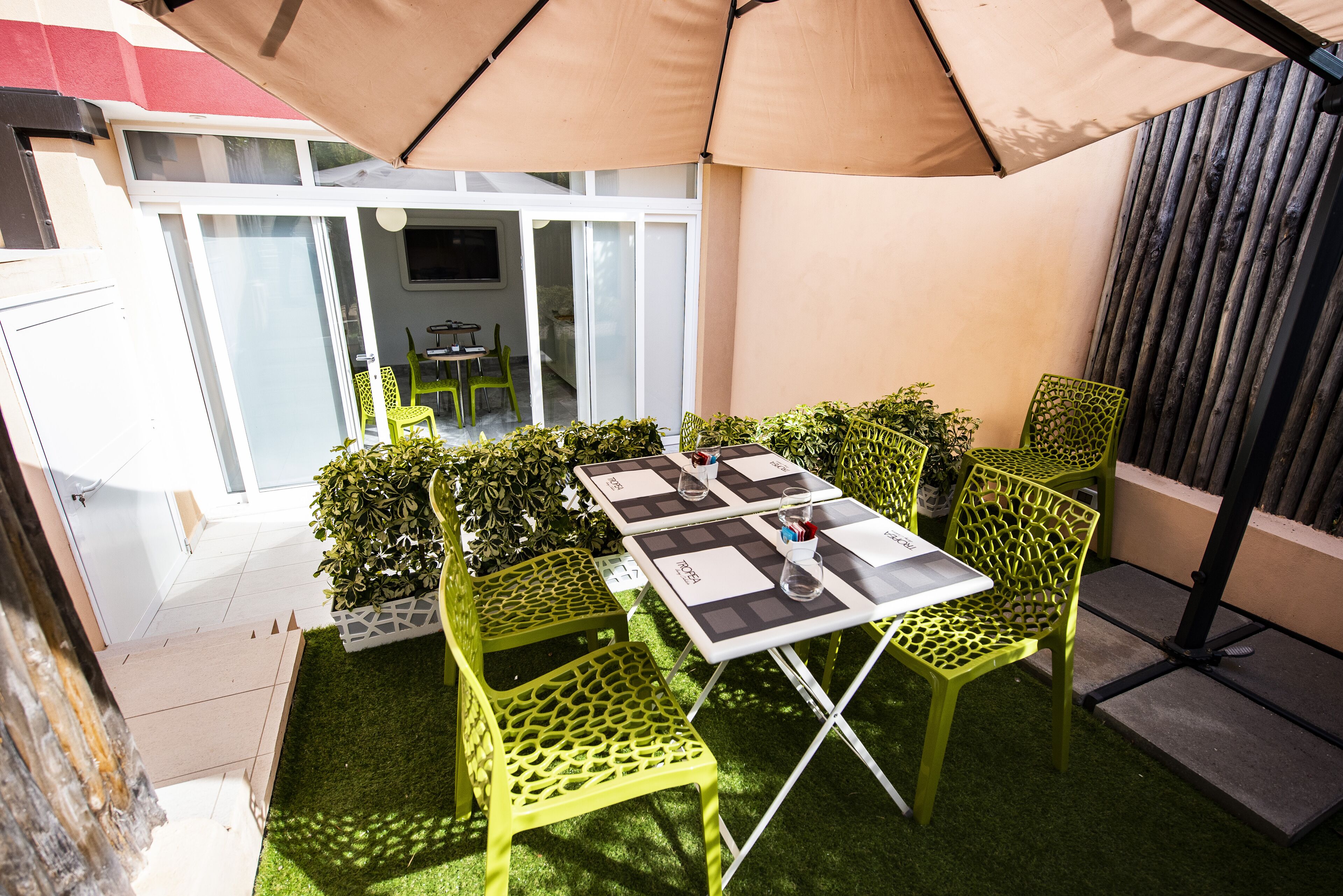 An outdoor patio with a table and chairs, surrounded by greenery and a large umbrella.