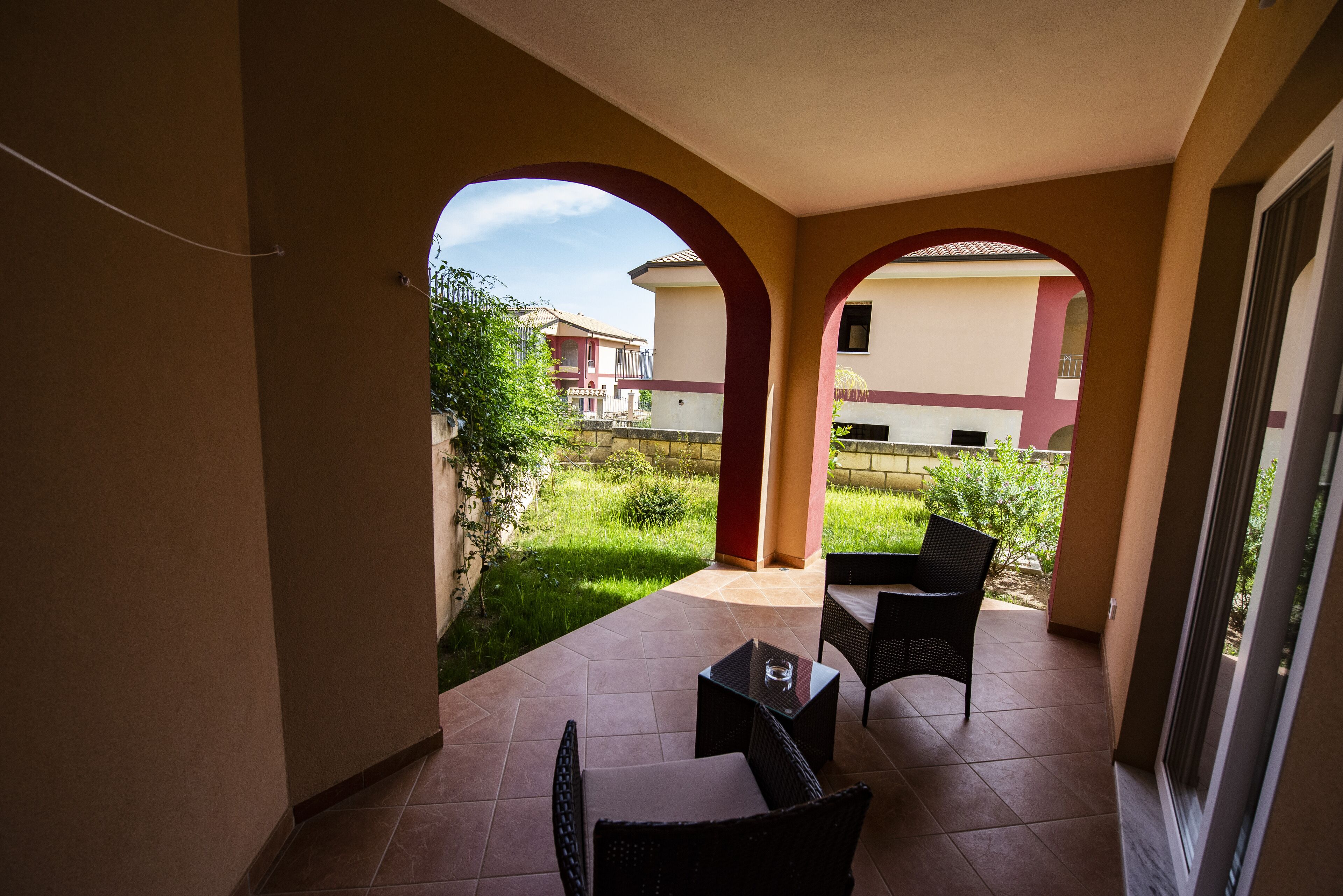 Portico esterno coperto con tavolo e sedie, vista su un giardino e un edificio sullo sfondo.