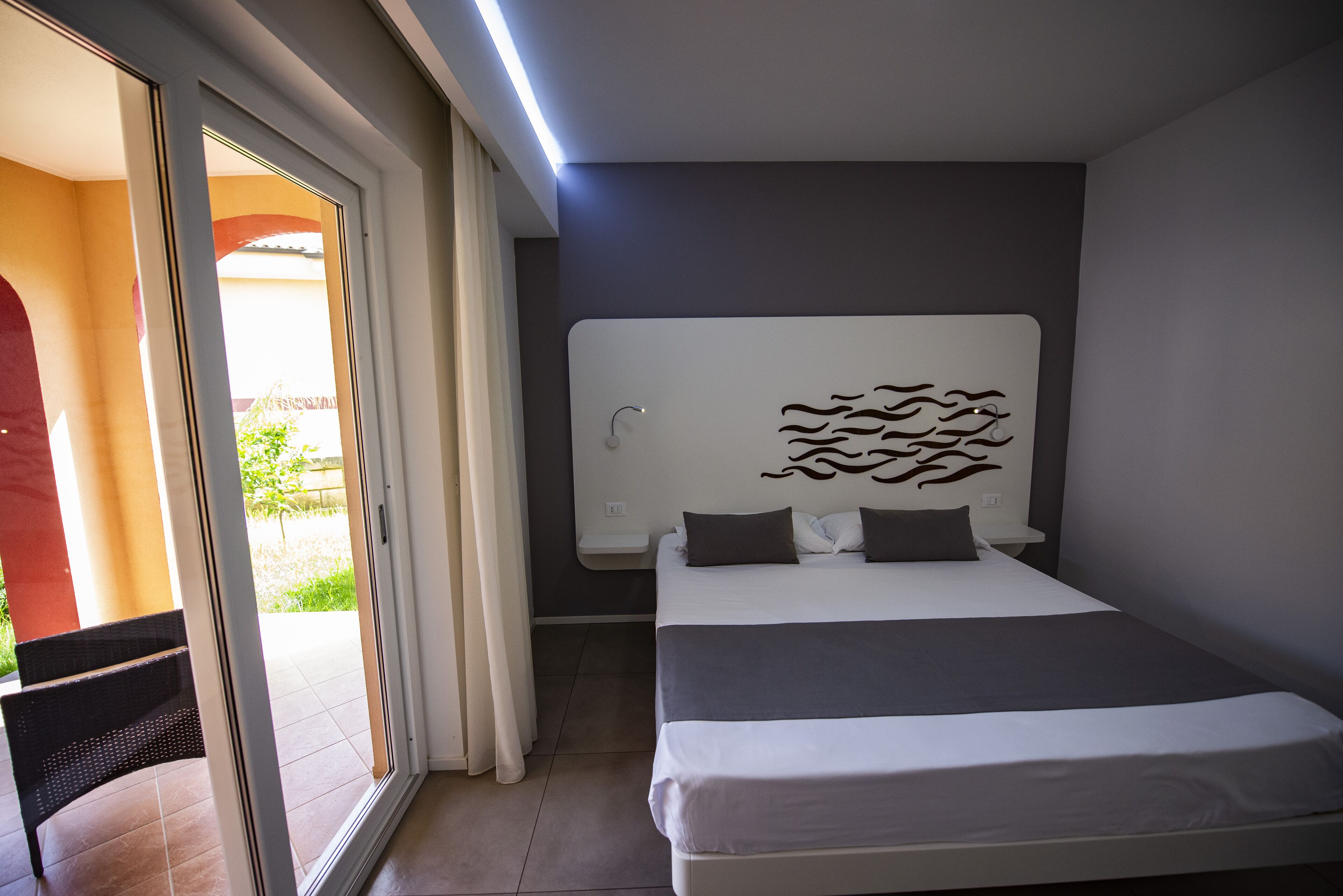 Una habitación de hotel moderna con una cama grande, cabecera con diseño de olas, puerta corredera de vidrio a un balcón y una mesita de noche pequeña.