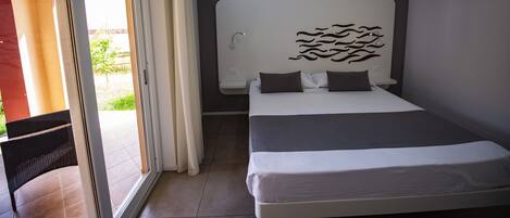 Una habitación de hotel moderna con una cama grande, cabecera con diseño de olas, puerta corredera de vidrio a un balcón y una mesita de noche pequeña.