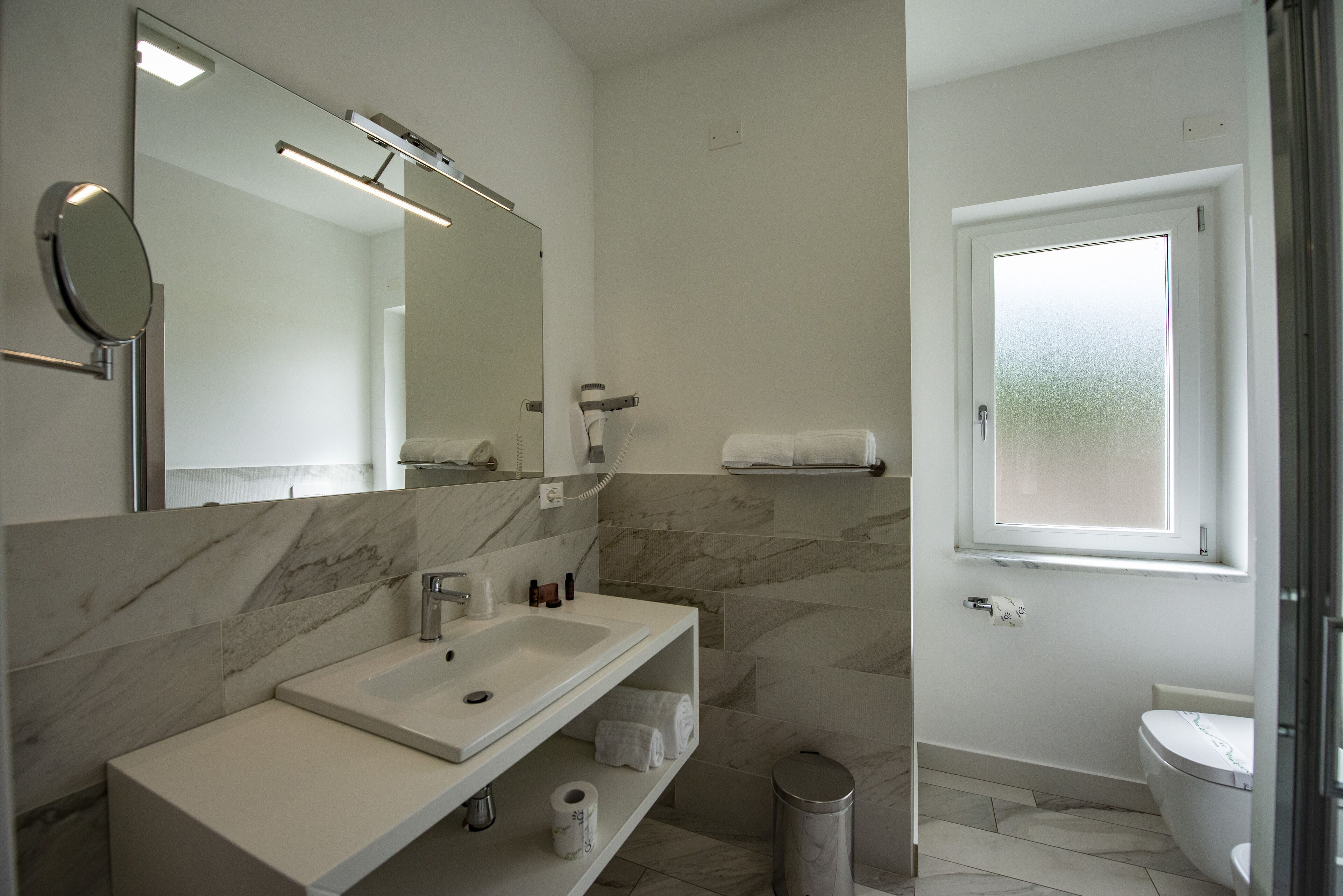 A modern bathroom with a marble vanity, a large mirror, a toilet, and a window.