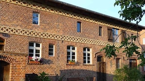 A brick building with a symmetrical facade, multiple windows, and a small garden area with a bench and potted plants. - Ferienwohnung Nitschke Zum Nusshof (Ahrensfelde)