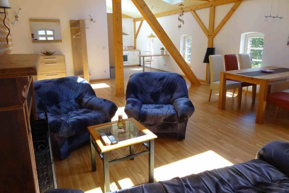 A spacious loft-style room with high ceilings and exposed wooden beams. It features two dark leather sofas, a glass coffee table, and a dining area with a wooden table and chairs.