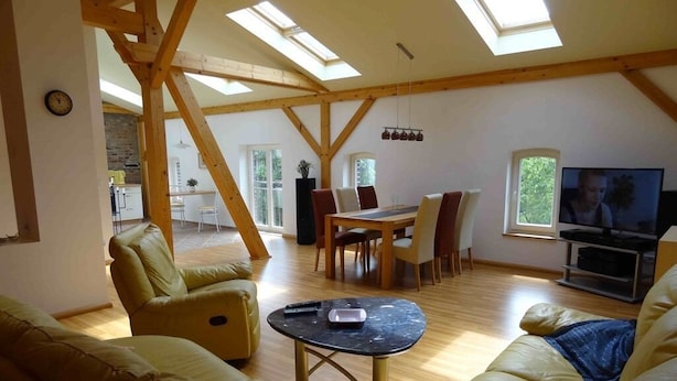A spacious living area with a high ceiling, wooden beams, a dining table with chairs, a sofa, and a television. - Ferienwohnung Nitschke Zum Nusshof (Ahrensfelde)