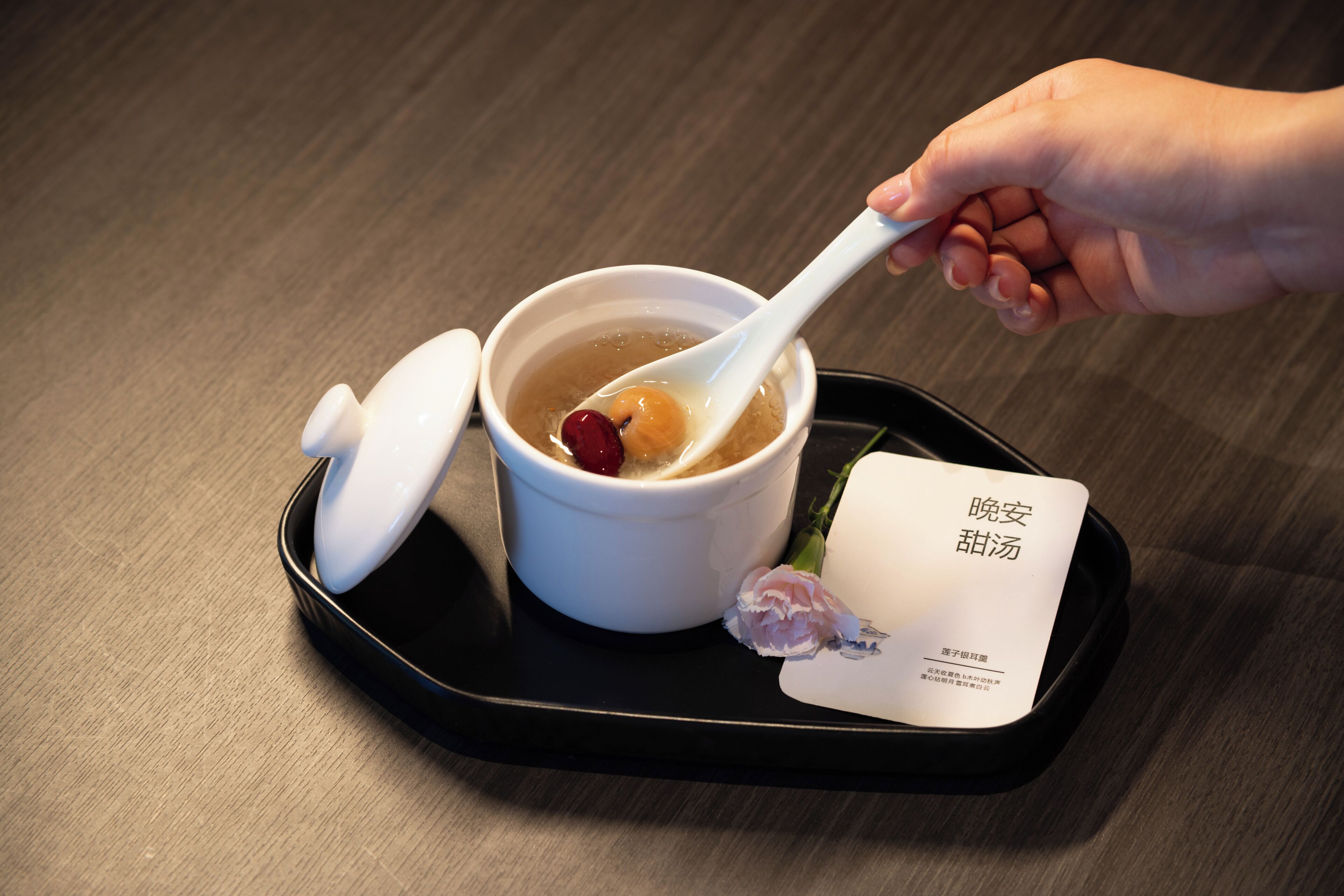A hand holding a spoon over a bowl of soup with a lid and a card beside it.
