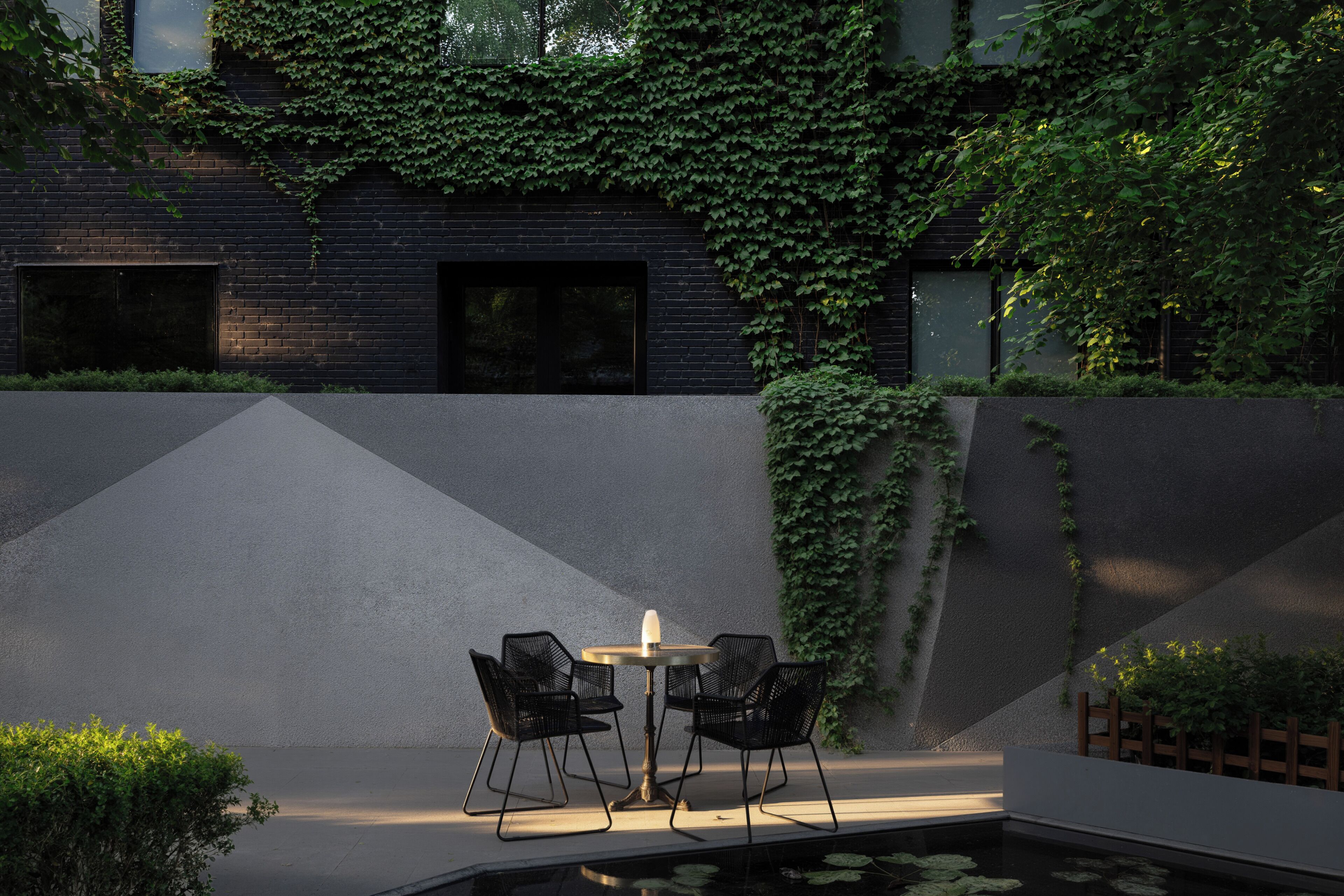 A modern outdoor seating area with a table and chairs, a small pool, and a building with ivy-covered walls.