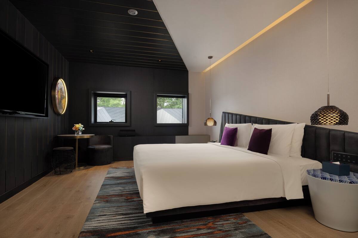 A modern hotel room with a large bed, a flat-screen TV, a small table with a vase, two windows, and a ceiling with a light fixture.