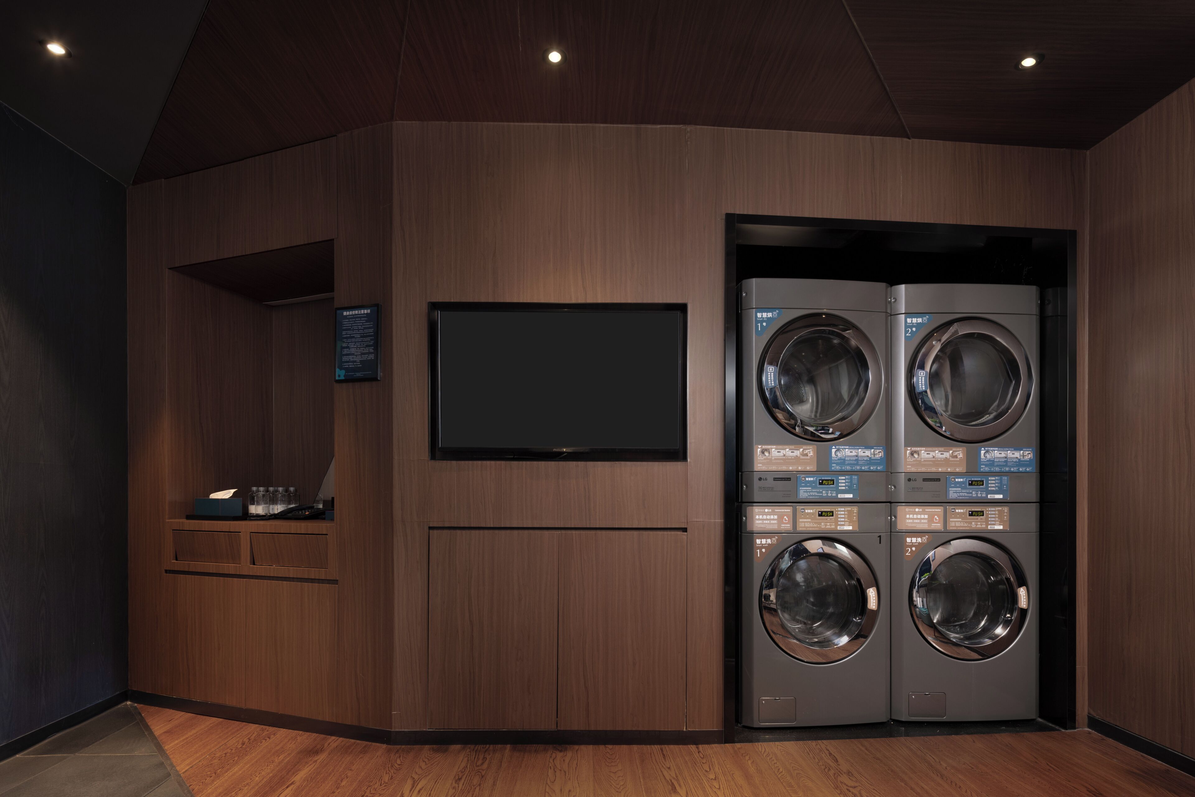 A laundry room with two stacked front-loading washing machines, a flat-screen TV mounted on the wall, and a small shelf with a tissue box and other items.