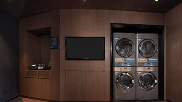 A laundry room with two stacked front-loading washing machines, a flat-screen TV mounted on the wall, and a small shelf with a tissue box and other items.