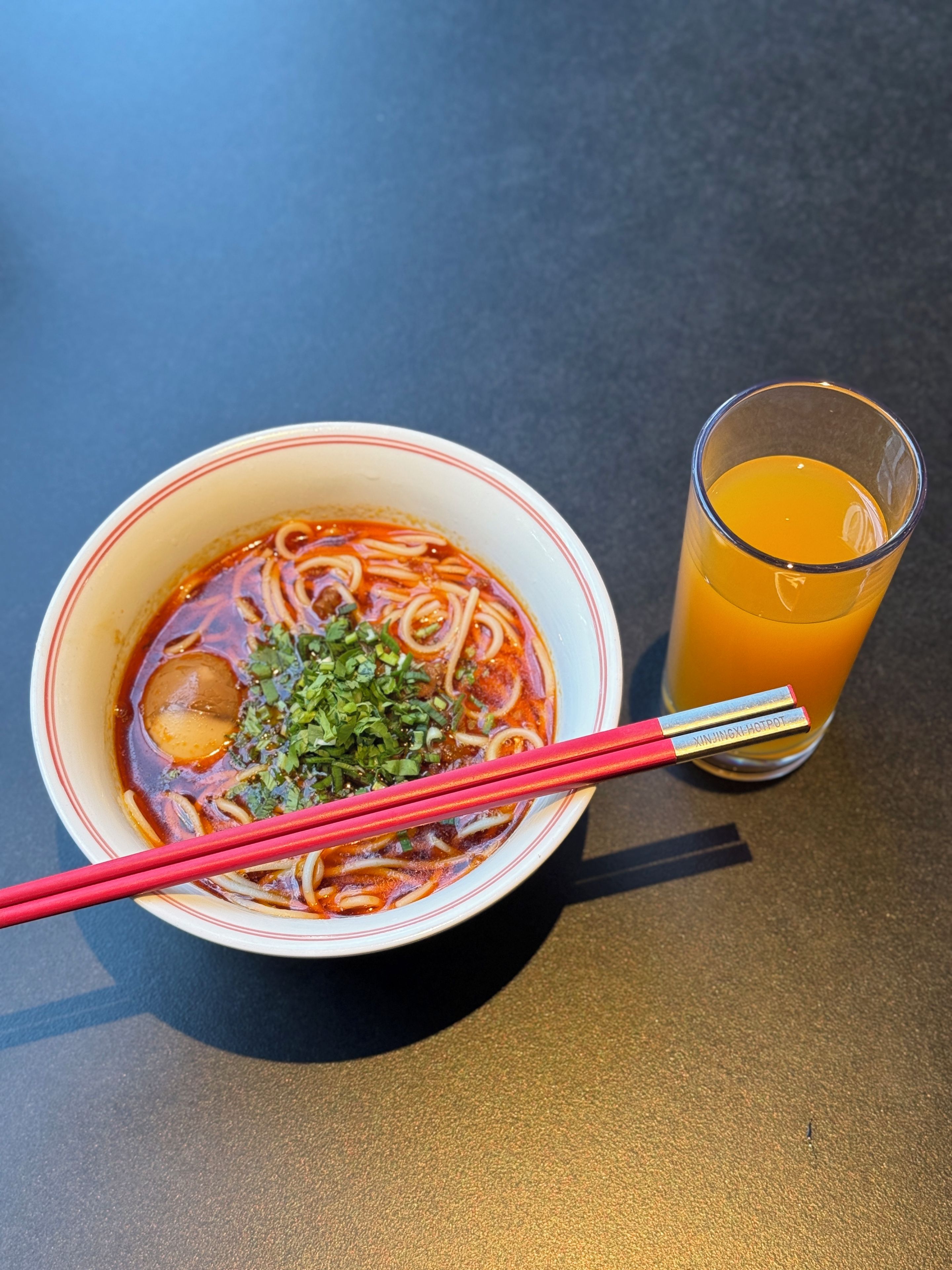 A bowl of ramen with chopsticks and a glass of orange juice on a table.