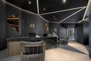 Un área de recepción moderna con mostrador de mármol, dos portátiles, una lámpara de escritorio y una zona de estar con una silla y un sofá.