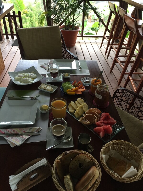 A dining table set with plates, cutlery, and various food items like fruits and bread, on an outdoor wooden deck with chairs and a potted plant.