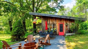 A rustic cabin with a red door, wooden exterior, and a patio with Adirondack chairs and a table.