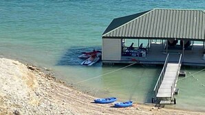 A dock with a covered seating area and a small building, boats on the shore, and calm water.