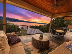 An outdoor patio with a view of a sunset over water, a round table with chairs, and a covered seating area. - Lakefront Home with Captained Part Boat, Private Dock, Beach, Game-room (Lago Vista)