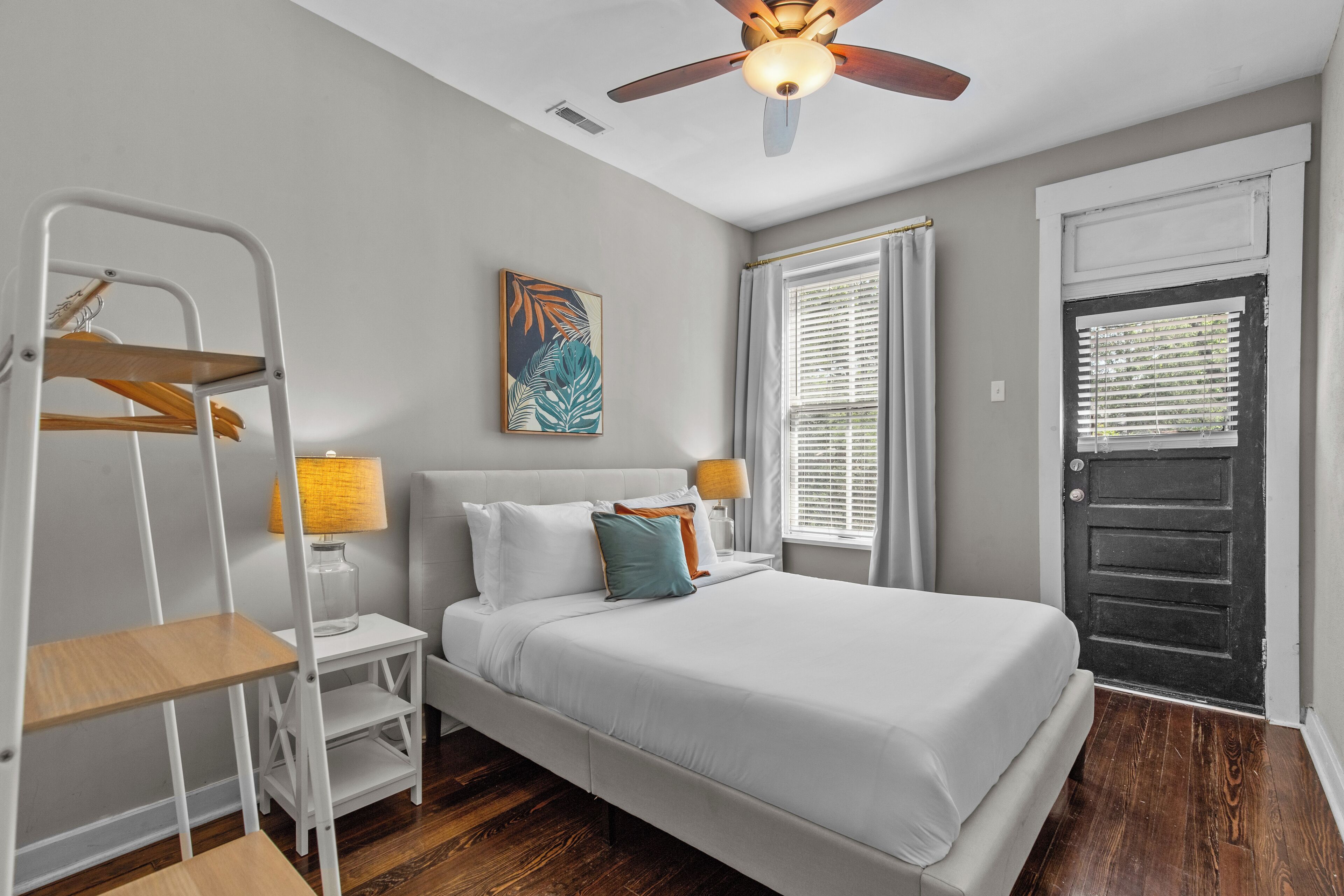 Une chambre à coucher comprenant un lit, un ventilateur de plafond, une fenêtre avec des stores, une porte et une petite table avec une lampe.