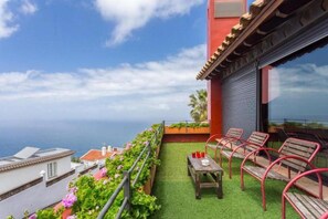 Un balcón con mesa y sillas, vista al mar, y un edificio de fachada roja.