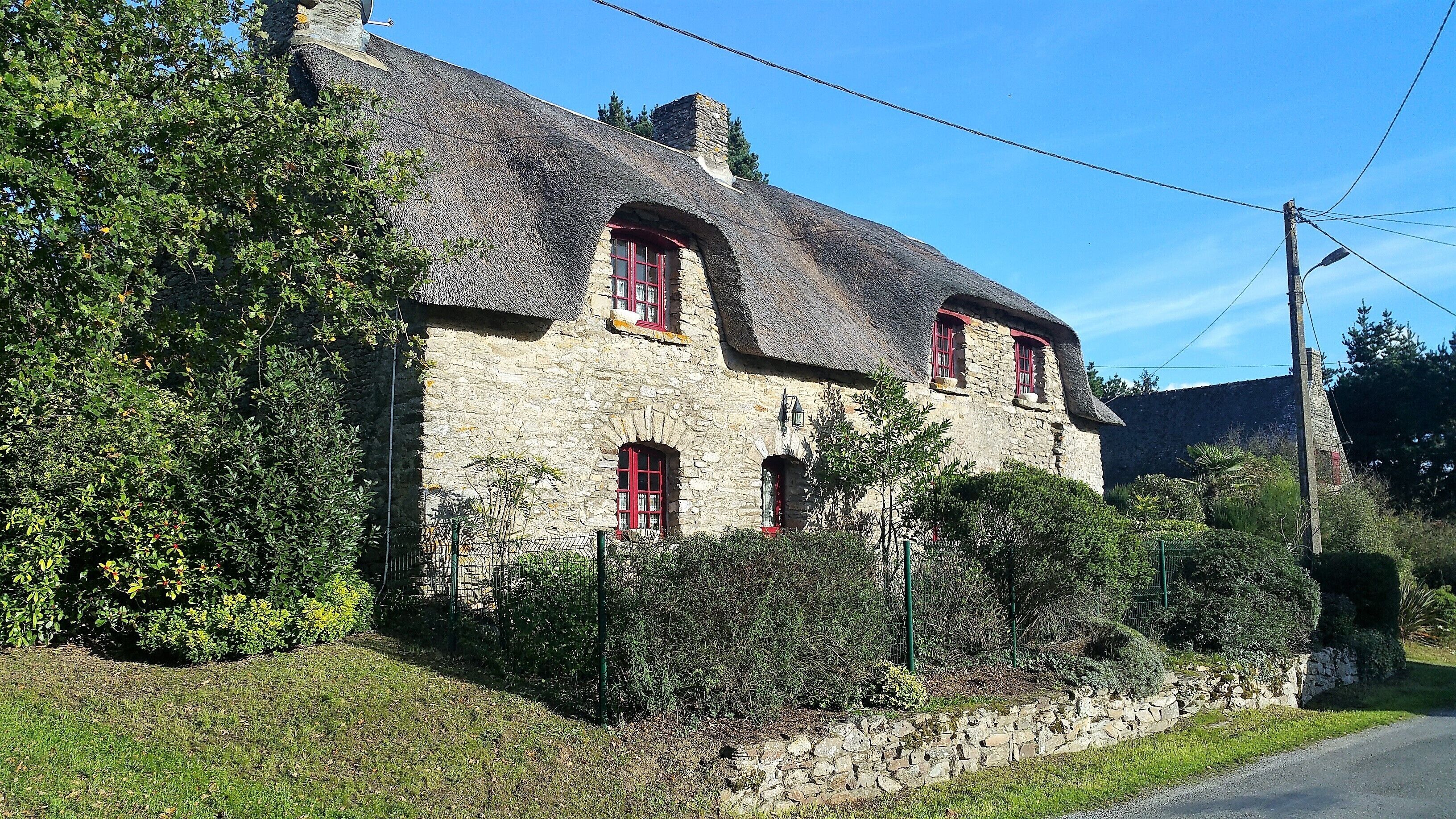 A stone house with a thatched roof, red windows, and surrounded by greenery.