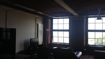 Um quarto com pouca luz, uma janela grande, uma televisão de tela plana e uma escrivaninha com um computador.