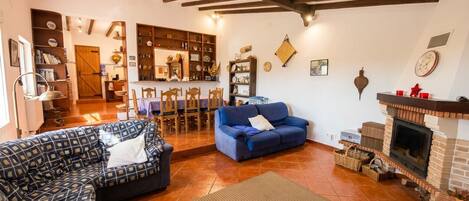 Uma sala de estar com sofá, poltrona azul, lareira e área de cozinha ao fundo.