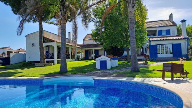 A poolside view of a residential property with a swimming pool, a covered patio, and a well-maintained lawn.