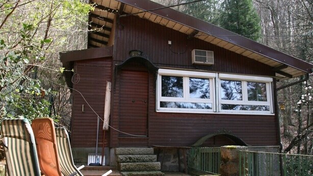 A wooden cabin with a large window, a porch with chairs, and a stone staircase leading to the entrance.