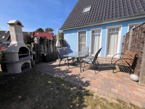 A backyard with a stone barbecue grill, a table with chairs, and a blue house with a tiled roof.
