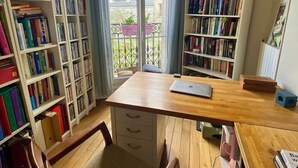 A study room with a wooden desk, bookshelves, a chair, and a view through a window. - Large Family Apartment in Paris (Paris)