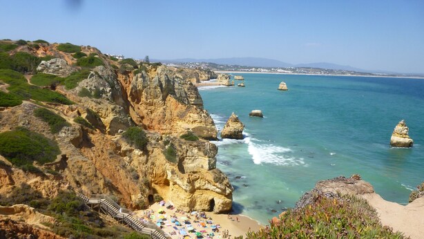 Paisatge costaner amb penya-segats rocosos, una platja amb ombrel·les i una vista clara del mar.