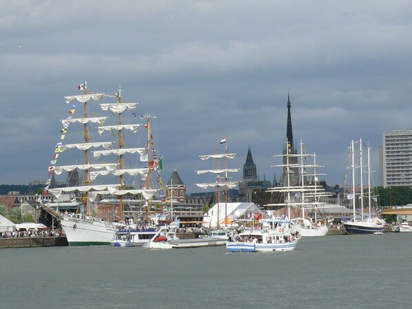 A harbor with multiple sailing ships docked, a prominent church spire in the background, and a modern building to the right.