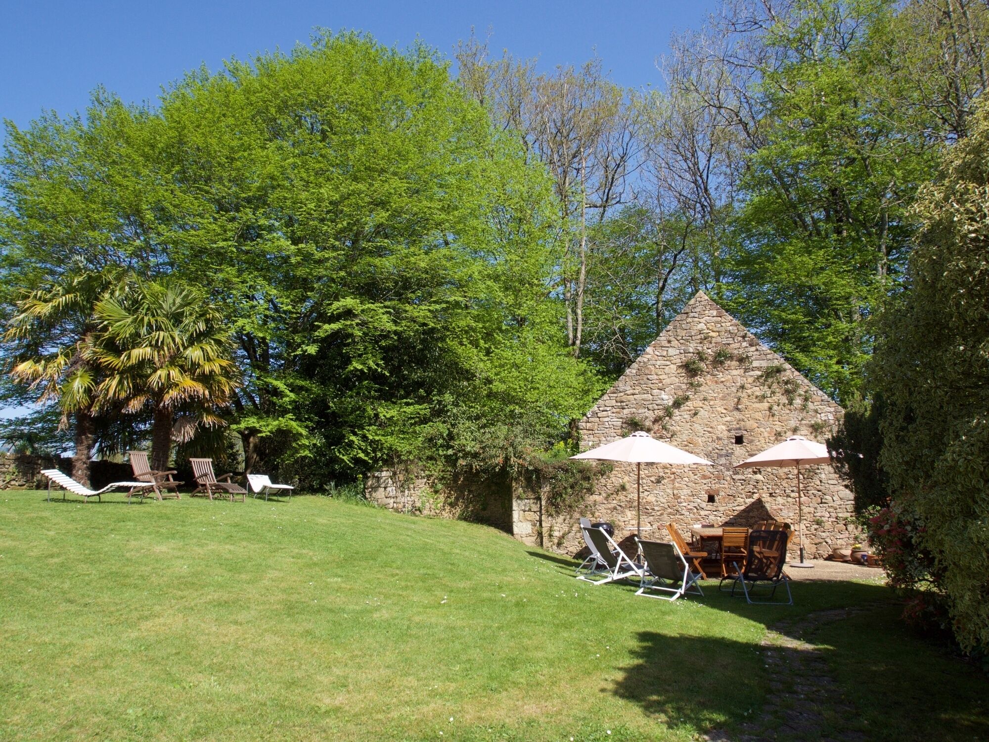 A stone building with a triangular roof, surrounded by lush green trees and a grassy lawn. There are lounge chairs and an umbrella providing shade.