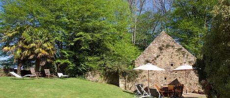 A stone building with a triangular roof, surrounded by lush green trees and a grassy lawn. There are lounge chairs and an umbrella providing shade.