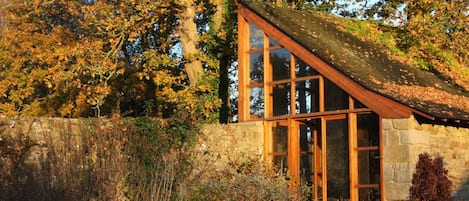 A stone building with large windows and a sloped roof, surrounded by autumn foliage.