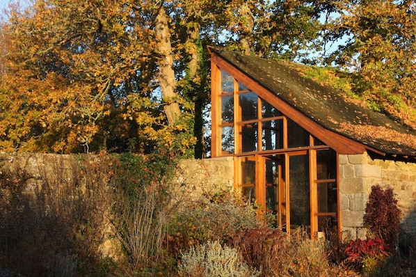 A stone building with large windows and a sloped roof, surrounded by autumn foliage.