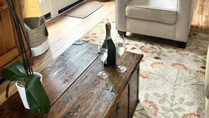 Sala de estar com poltrona bege, mesa de centro de madeira, uma garrafa de champanhe e duas taças. Há um espelho na parede, um vaso com flores e uma porta que dá para o exterior.